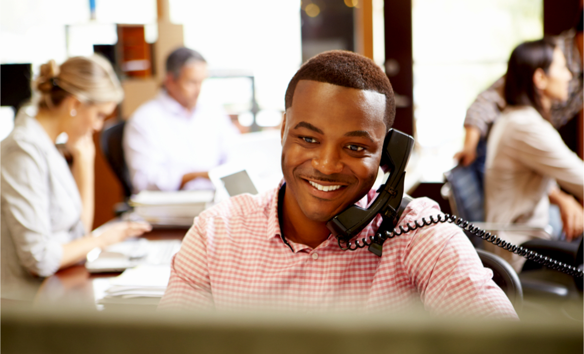 A smiling business man sits at his desk making a phone call to a client
