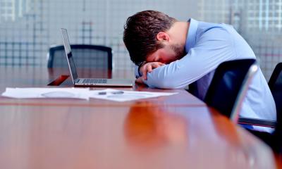 Man in blue shirt with his head in his arms in font of a laptop