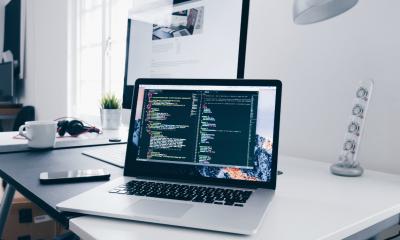 Laptop on a desk showing coding used to build apps with a computer in the background