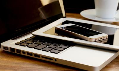 A laptop, tablet, and phone resting on top of a wooden desk with a white mug in the background