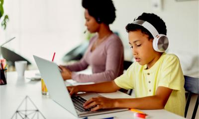 A boy completes his school work while his mother works remotely in the background