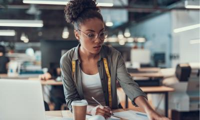 Woman wearing glasses stood at a desk holding a pen with a coffee in front of her