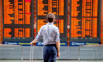 A young business man stands in front of a airport departures board