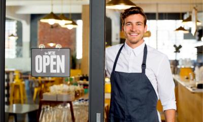 Portrait of smiling owner standing at his restaurant gate with open signboard.