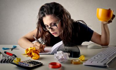 Woman wearing glasses sat at a messy desk holding a yellow mug in the air