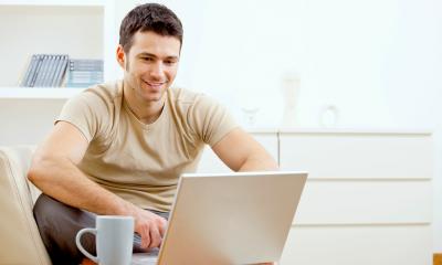 Man in a beige t-shirt working from home on his laptop with a mug beside him